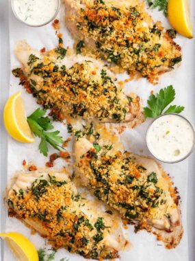 Golden baked fish fillets topped with a crispy herb breadcrumb crust, served with lemon wedges and dipping sauce on a parchment-lined tray.