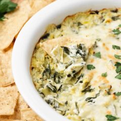 Baked spinach artichoke dip in a white bakish dish with a tortilla chip dipped into it to show cheesy, creamy texture.