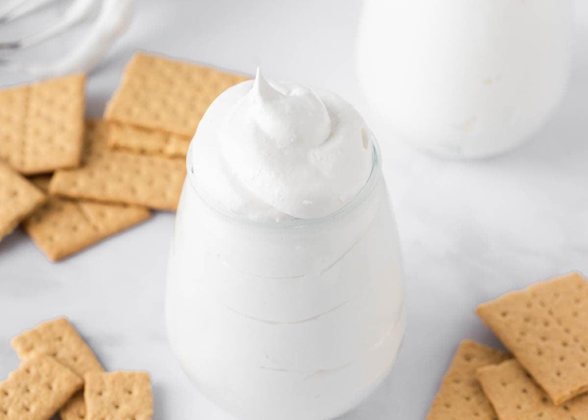 Homemade marshmallow fluff transforms sweet treats into fluffy, cloud-like confections. Perfect for topping desserts, it's far superior to store-bought marshmallow fluff. Fluffy homemade marshmallow fluff with soft peaks in a glass jar served with graham crackers for dipping.