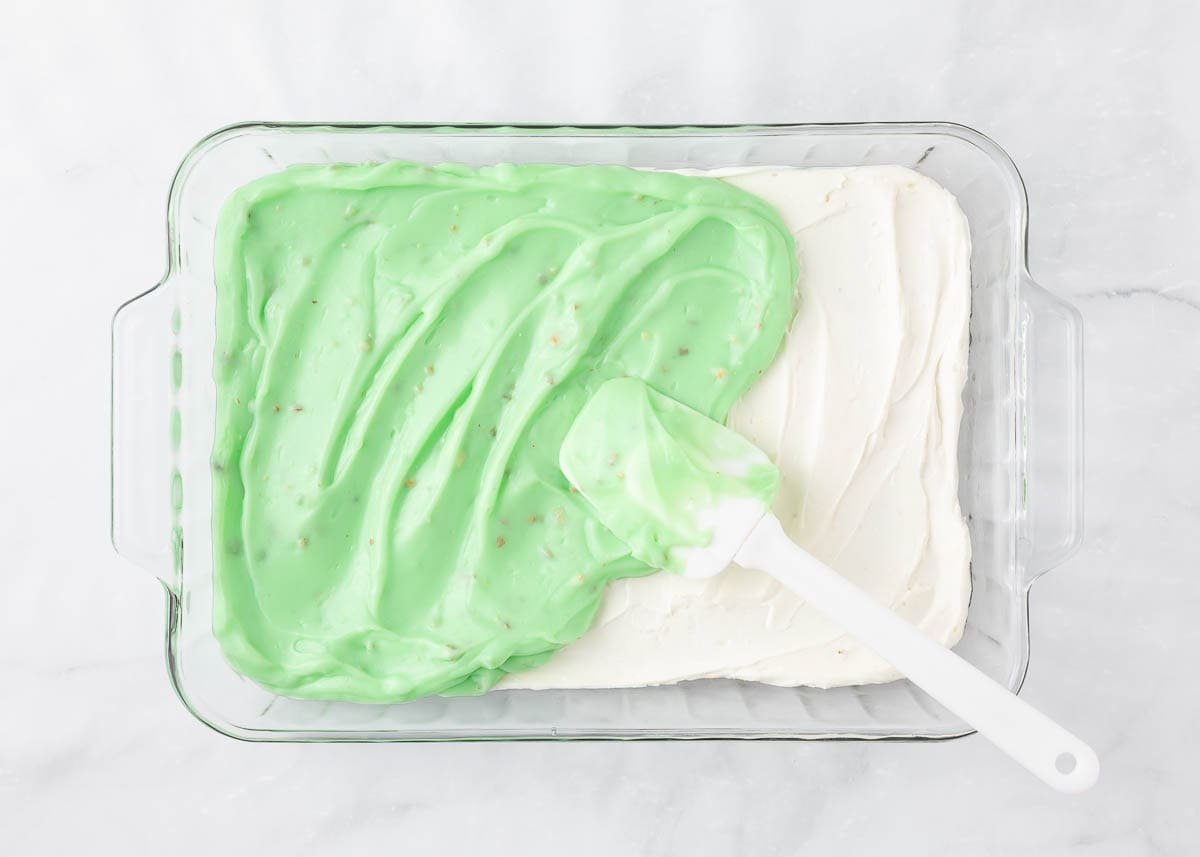 Pistachio pudding being spread over cheesecake layer in a baking dish.