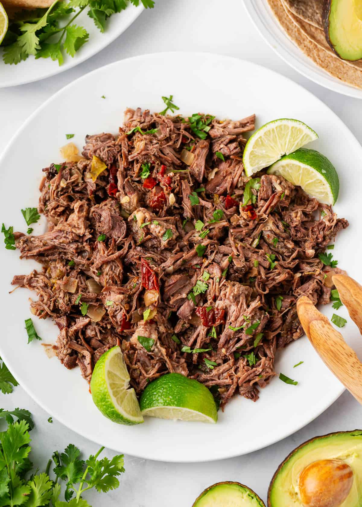 This slow cooker beef barbacoa is a Mexican staple easy to make in the crockpot! Cooking low and slow results in tender beef full of flavorful spices ready to use in tacos, burritos or bowls! Barbacoa beef shredded on a plate with lime wedges.
