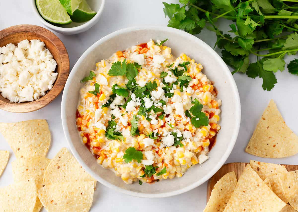 Elote dip in a white bowl topped with queso fresco cheese and cilantro, surrounded by tortilla chips and fresh limes.