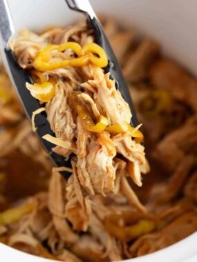 Tongs lifting a serving of shredded Mississippi chicken with pepperoncini peppers from a white crockpot.