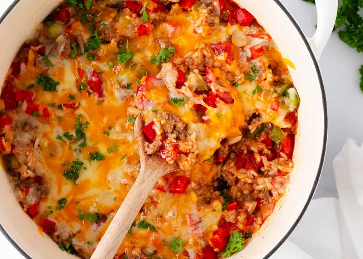 This stuffed pepper casserole brings all the cozy vibes of the classic dish but cuts down on prep and cooking time, making it a perfect weeknight meal. Close-up of stuffed pepper casserole in a white pot, with melted cheddar over ground beef, rice and red and green bell peppers in tomato sauce, topped with fresh parsley, a wooden spoon scooping a serving.