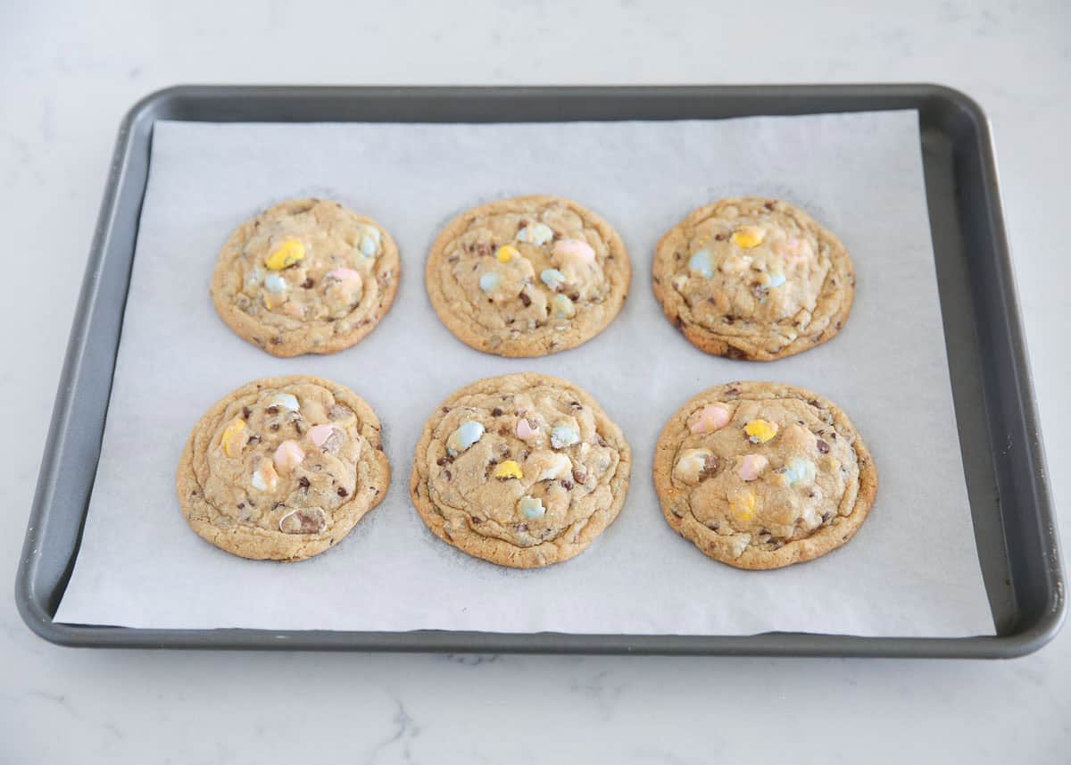 Six baked Cadbury egg cookies with pastel mini eggs and mini chocolate chips on a parchment-lined baking sheet.