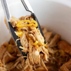 Tongs lifting a serving of shredded Mississippi chicken with pepperoncini peppers and au jus from a white crockpot.