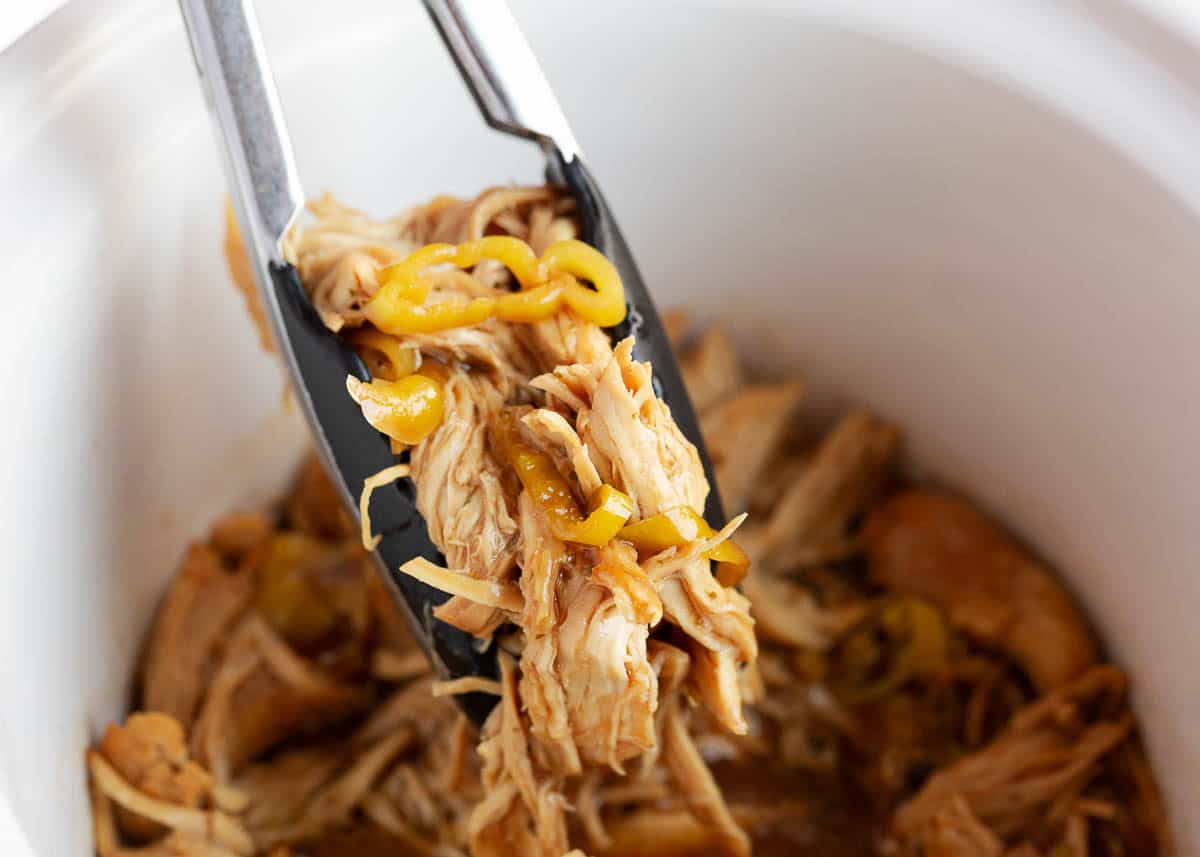 Tongs lifting a serving of shredded Mississippi chicken with pepperoncini peppers and au jus from a white crockpot.