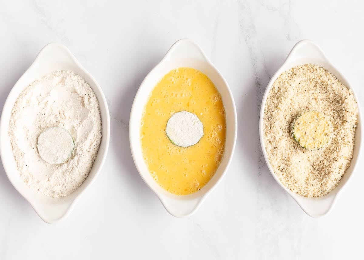 Three white oval dishes lined up with flour, beaten egg, and seasoned panko breadcrumbs, with a zucchini round being dredged in each.