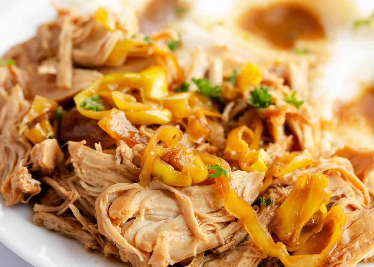 Close-up of shredded Mississippi chicken topped with sliced pepperoncini peppers and parsley, served next to mashed potatoes.