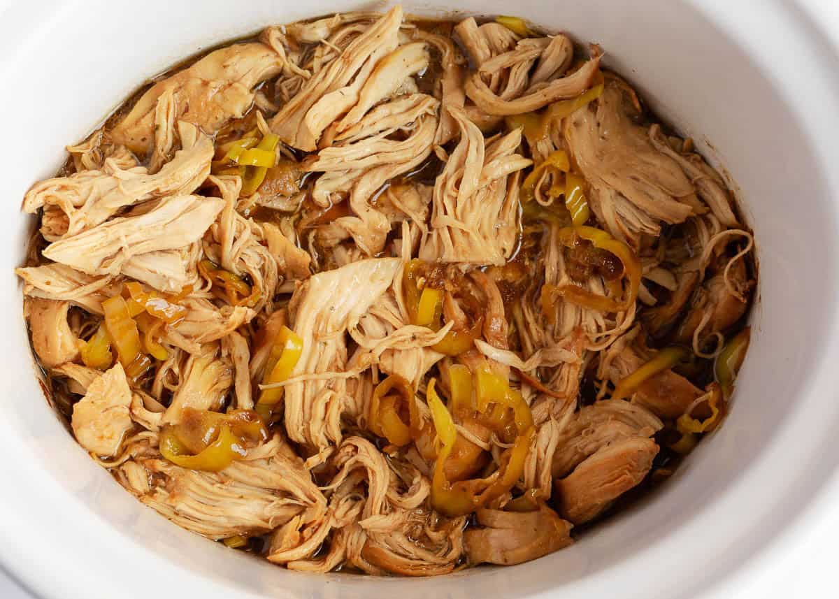 Cooked Mississippi chicken in a white crockpot with pepperoncini peppers and au jus sauce, ready to be shredded.