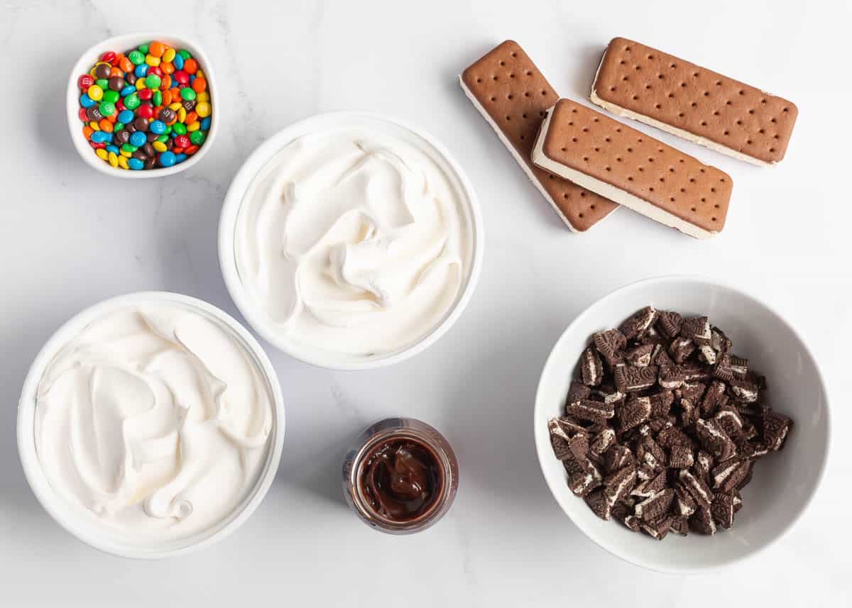 This ice cream sandwich cake is the best summer dessert! It's easy to make this no-bake treat with ice cream sandwiches, hot fudge sauce, Oreo's and Cool Whip. Whip cream, ice cream sandwiches, Oreos, hot fudge and M&Ms on the counter.