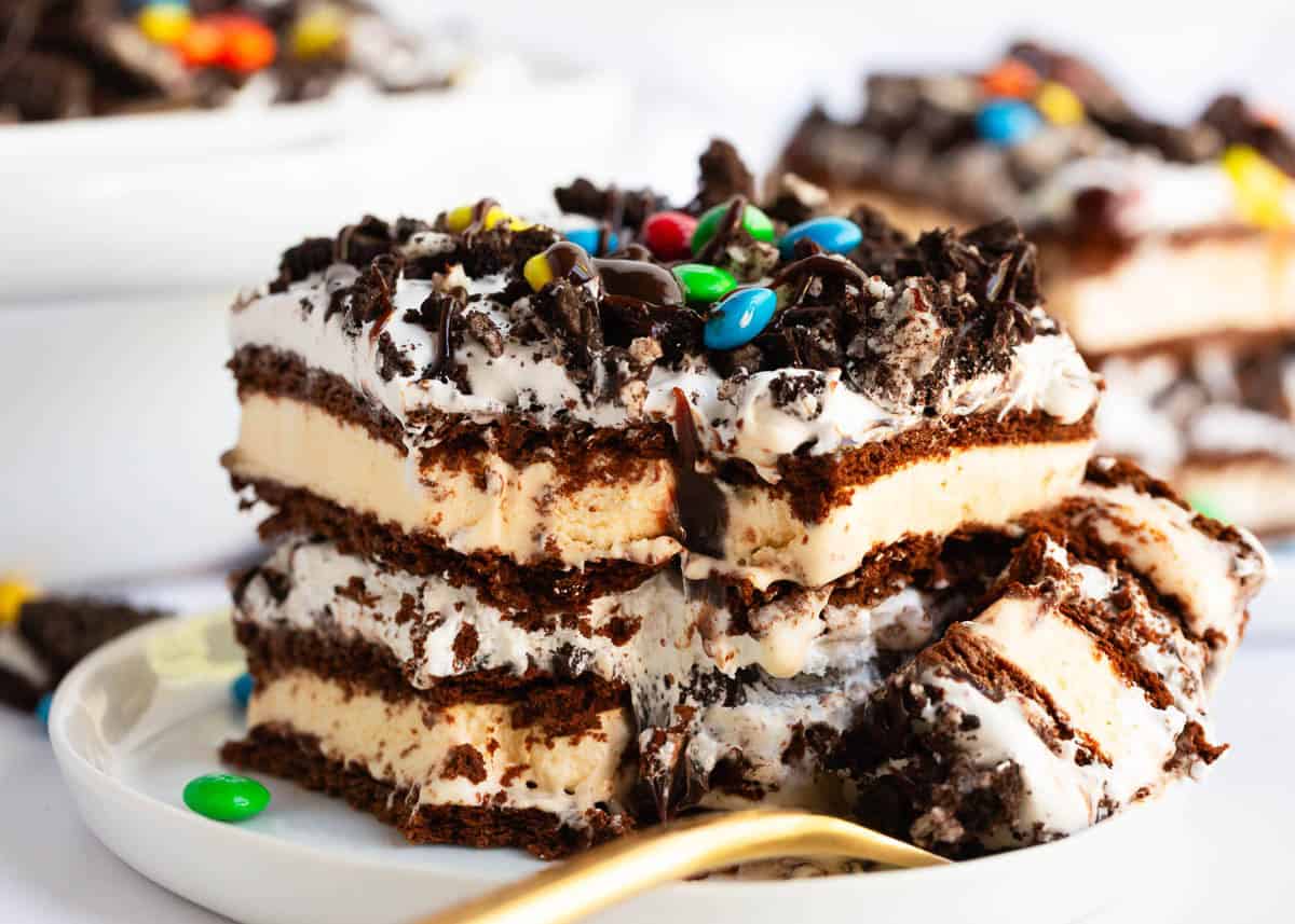This ice cream sandwich cake is the best summer dessert! It's easy to make this no-bake treat with ice cream sandwiches, hot fudge sauce, Oreo's and Cool Whip. Slice of ice cream sandwich cake on a white plate with M&Ms, Oreo crumbles and hot fudge.