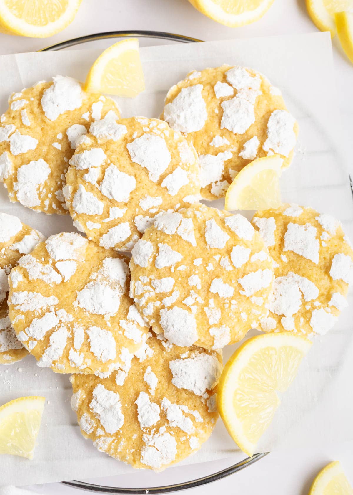 Thick and chewy lemon crinkle cookies have the perfect balance of sweet and buttery lemon flavor. They're rolled in powdered sugar and baked until the surface cracks! Lemon crinkle cookies on parchment covered cooling rack with fresh lemon slices.