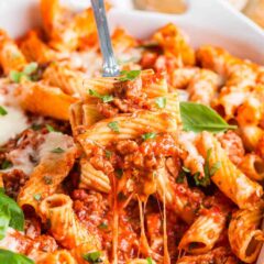 Close-up of cheesy baked rigatoni with meat sauce in a white baking dish. A fork lifts a serving of rigatoni showing melted mozzarella cheese stretching with fresh basil garnish.