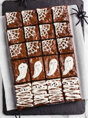 Tray of fudgy Halloween brownies decorated with melted white chocolate spider webs, ghost faces, and mummy designs with candy eyes.