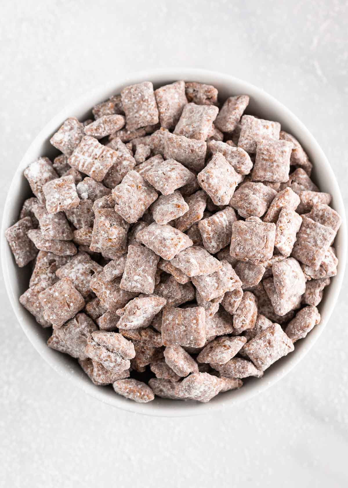 Chocolate and peanut butter coated cereal dusted in powdered sugar makes this puppy chow recipe irresistible. This snack is perfect for a last minute dessert! Puppy chow in a white bowl.
