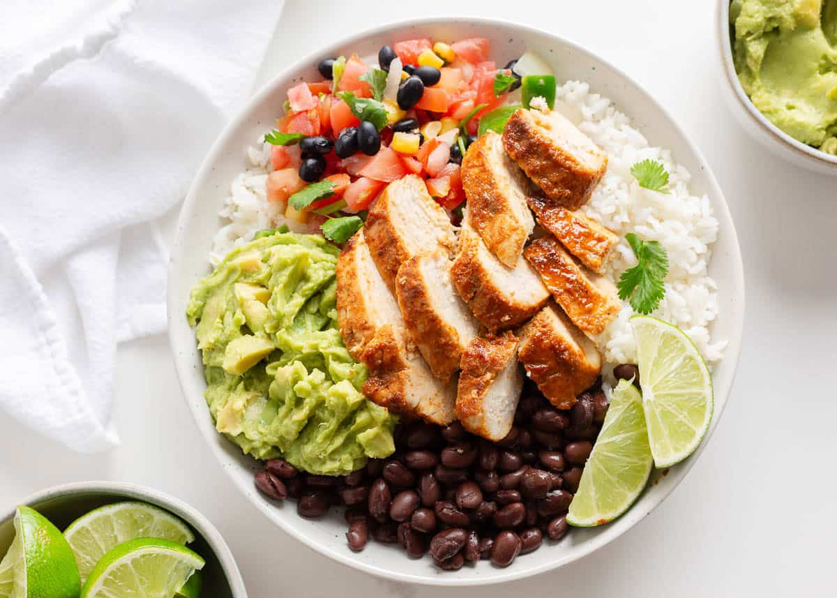 Skip ordering out and make copycat Chipotle chicken burrito bowls at home. Easy to assemble with seasoned chicken breast, rice, beans and toppings for dinner or meal prep! Burrito bowl topped with sliced chipotle chicken, creamy guacamole, black beans, pico de gallo, and lime wedges over white rice.