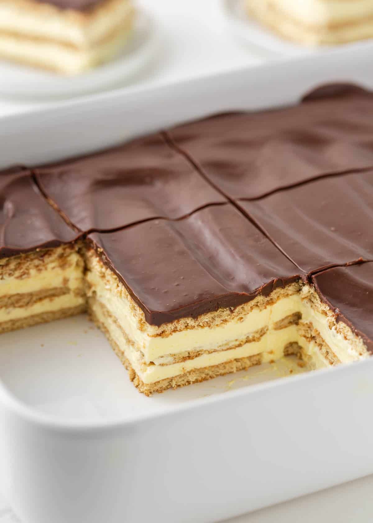 This chocolate eclair cake is an easy no-bake dessert made with layers of graham crackers separated by thick luscious cream and topped with a chocolate frosting. Eclair cake with chocolate ganache sliced in baking dish revealing layered graham crackers and vanilla filling.