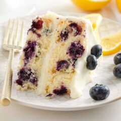 A slice of lemon blueberry layer cake on a plate garnished with fresh lemon wedges and blueberries.