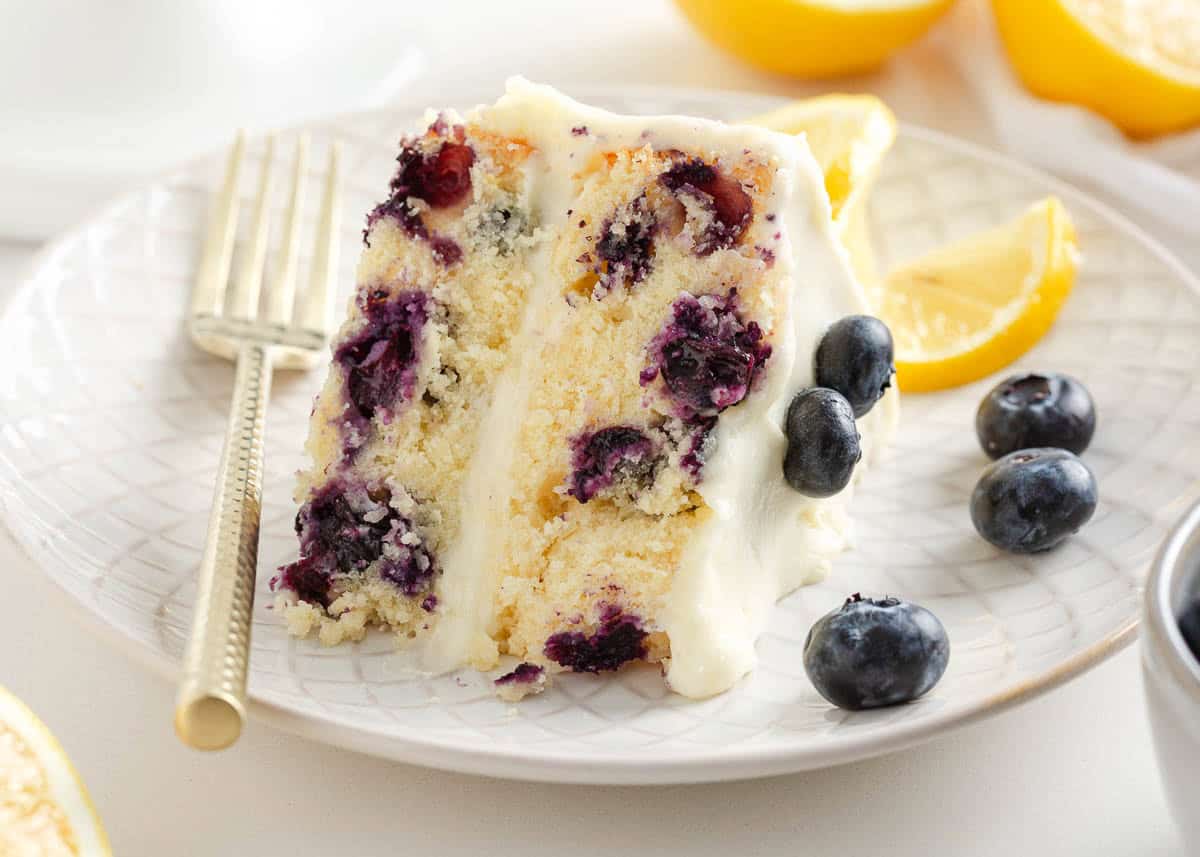 This lemon blueberry cake has a soft and tender crumb flecked with juicy blueberries and a lemon cream cheese frosting delivers the ultimate balance of sweet and tart! A slice of lemon blueberry cake on a plate.