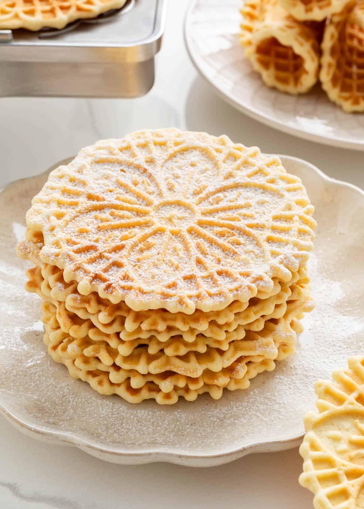 Pizzelle cookies stacked on a white counter dusted with powdered sugar.
