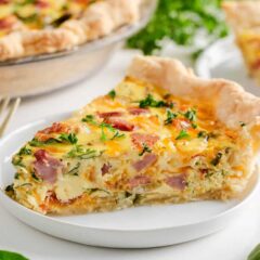 A slice of ham and cheese quiche on a plate showing flaky crust, creamy egg filling and ham pieces.