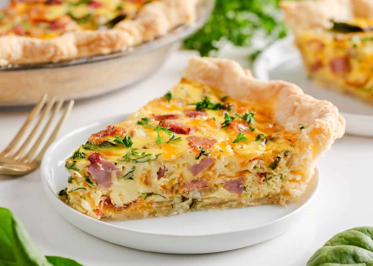This easy ham and cheese quiche is made with a buttery, flaky pie crust base and rich, cheesy egg filling that is perfect for serving at breakfast, brunch, or holiday mornings! You will love the savory and comforting flavors blended together! A slice of ham and cheese quiche on a plate.