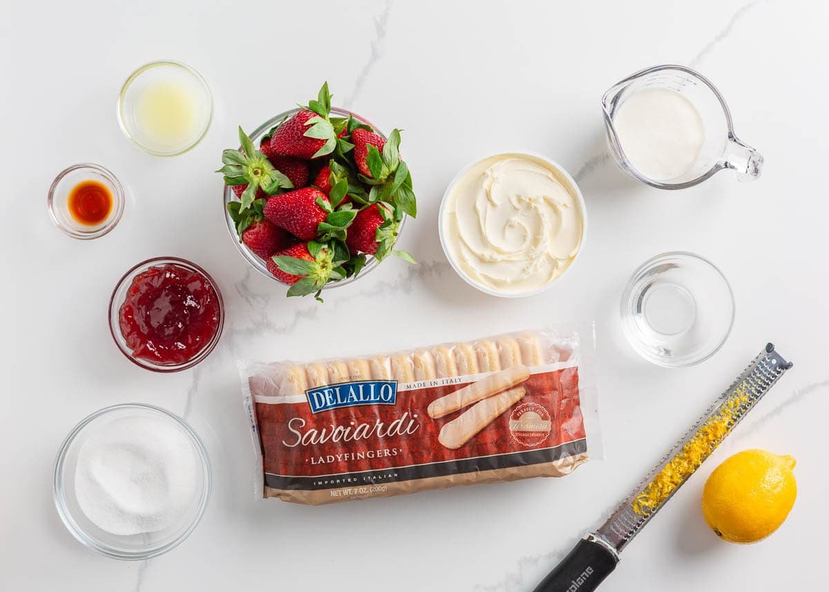 Strawberries, ladyfinger cookies, strawberry jam, mascarpone cheese, whipped cream and lemon on the counter.