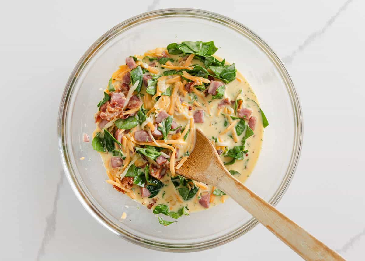 Bacon, ham, eggs, spinach, cheese and milk being stirred together in a bowl.