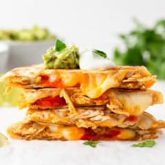 Shredded chicken quesadillas stacked on a plate with melted cheese, sautéed peppers and onions, topped with guacamole and sour cream.