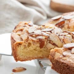 Close up of an almond torte with a slice removed, topped with sliced almonds and a dusting of powdered sugar.
