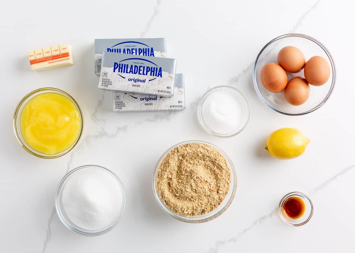 Lemon, lemon curd, cream cheese, eggs, sugar, graham cracker crumbs and butter on the counter.