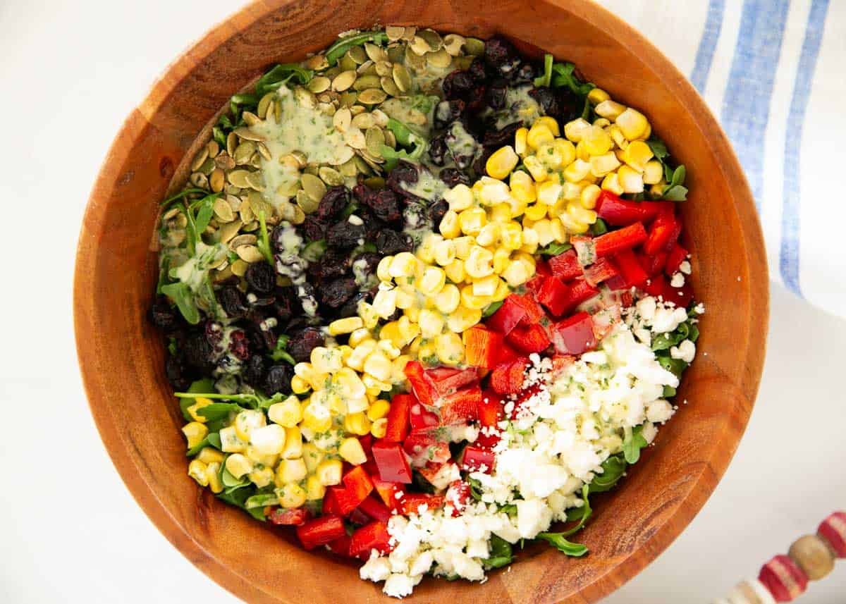 This arugula chopped salad is a copycat of the Wildflower and Gladly restaurant favorite made with arugula, red peppers, corn, feta, cranberries, and pumpkin seeds in a creamy basil dressing. Arugula chopped salad in a bowl with corn, red pepper, cranberries, sunflower seeds and feta.