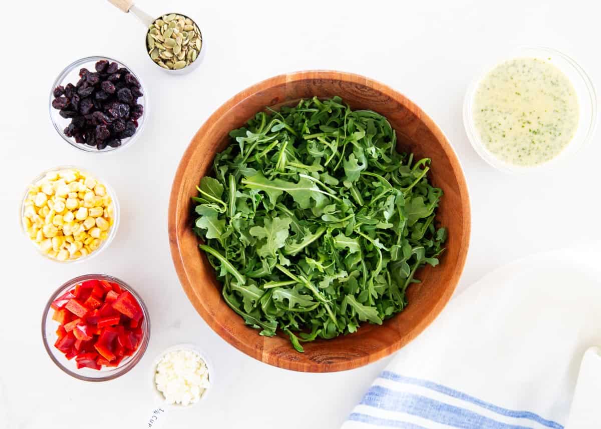 Arugula, corn, diced red pepper, dried cranberries, pumpkin seeds, creamy basil dressing and feta cheese on a counter.