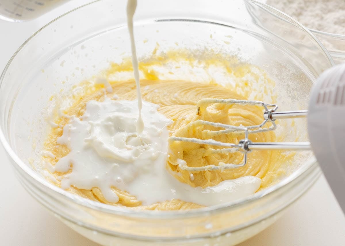 Wet ingredients being poured into lemon cake batter while mixing for lemon blueberry layer cake.