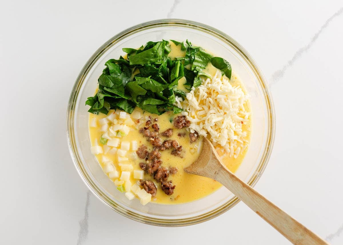 Mixing eggs, diced O’Brien potatoes with onions and peppers, spinach, sausage and cheese for Easy Potatoes O’Brien breakfast casserole.