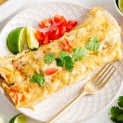 Cream cheese chicken enchilada topped with melted cheese, cilantro, and diced tomatoes, served with lime wedges.