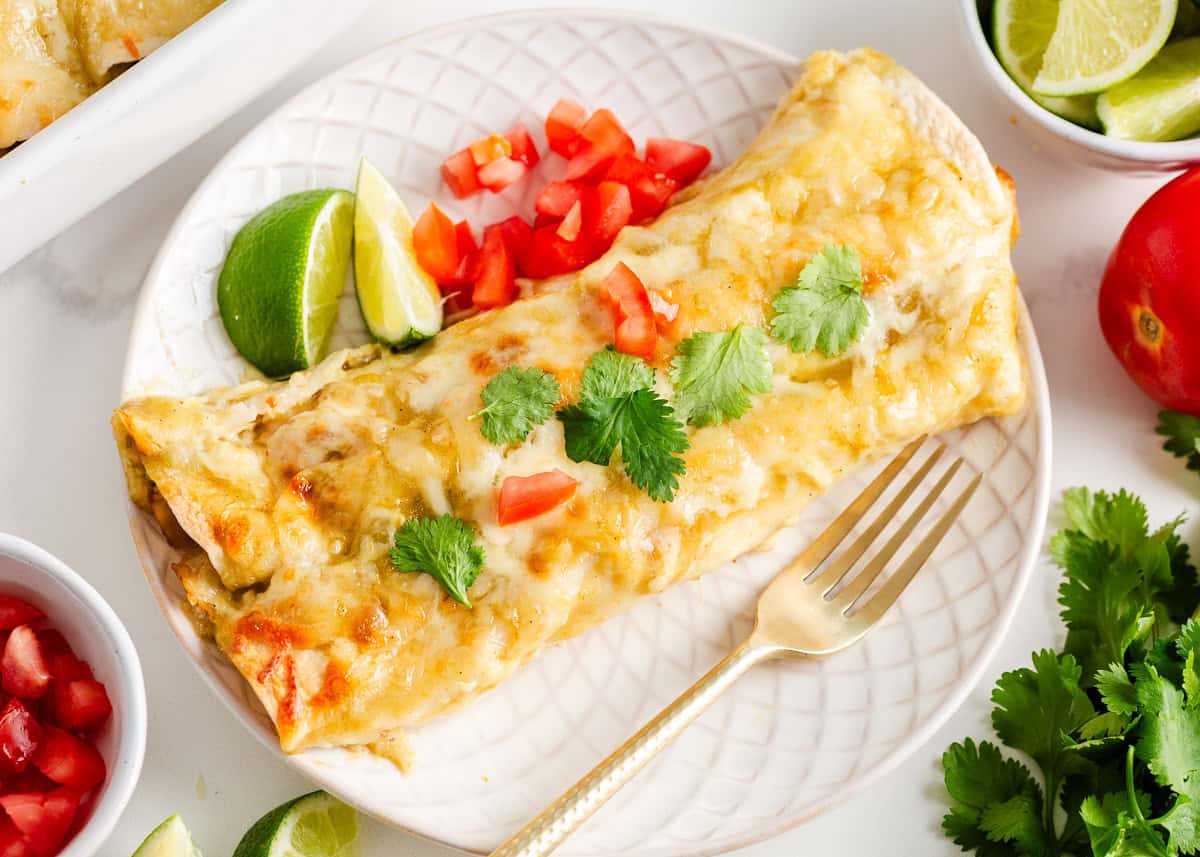 Cream cheese chicken enchilada topped with melted cheese, cilantro, and diced tomatoes, served with lime wedges.