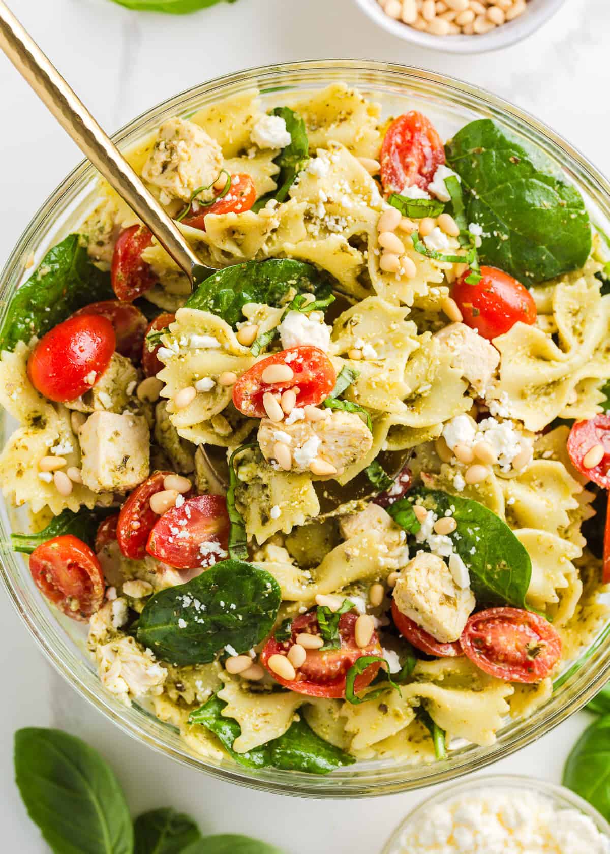 Colorful chicken pesto pasta salad served in a large clear bowl with serving spoon.