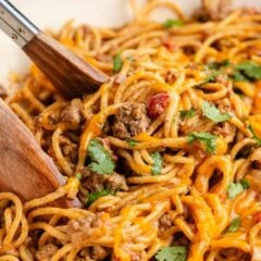 Serving tongs lifting a cheesy portion of taco spaghetti with ground beef.