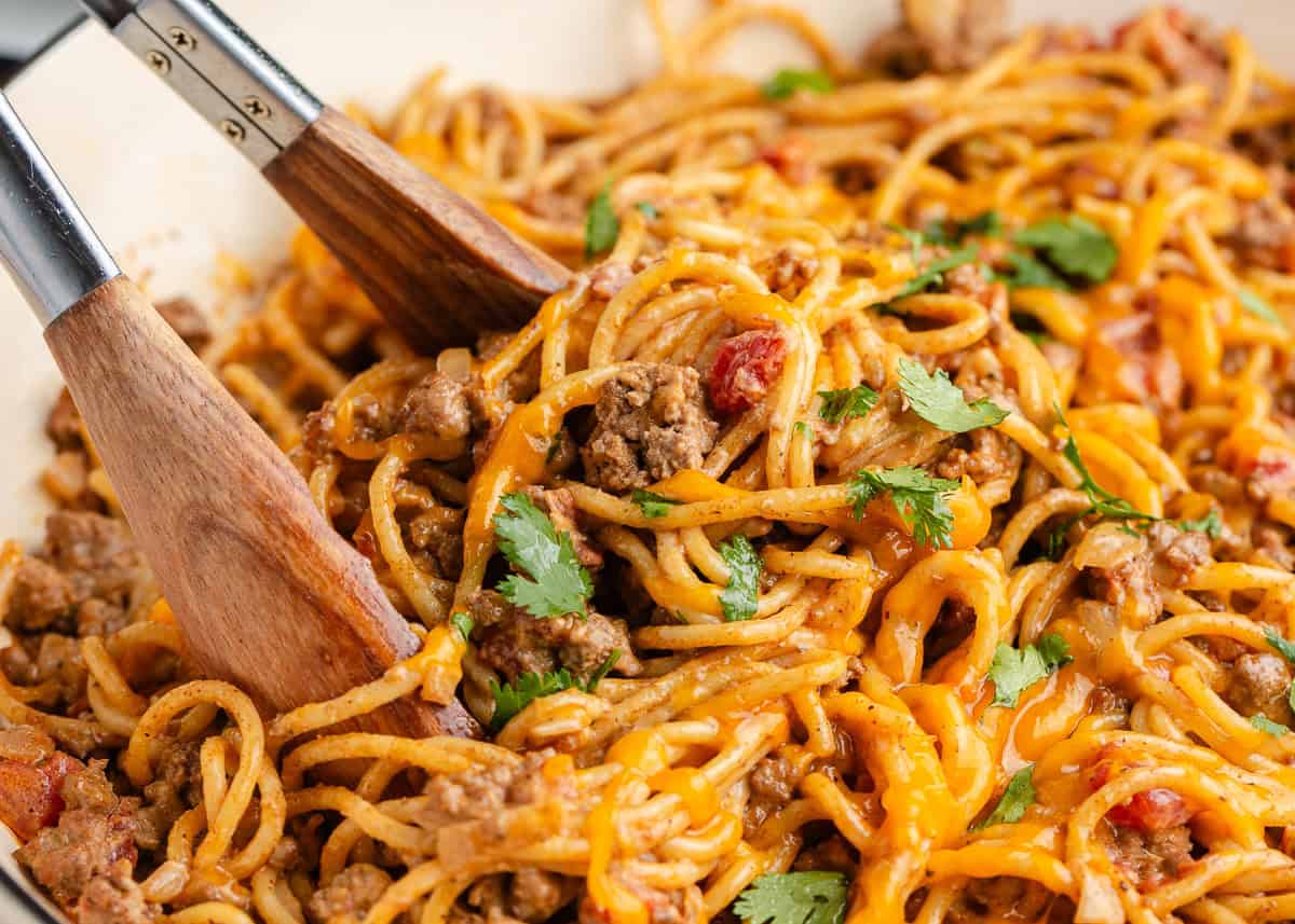 This taco spaghetti combines two favorites in one creamy, cheesy, zesty pasta dish. It's perfect for a quick weeknight dinner! Serving tongs lifting a cheesy portion of taco spaghetti with ground beef.