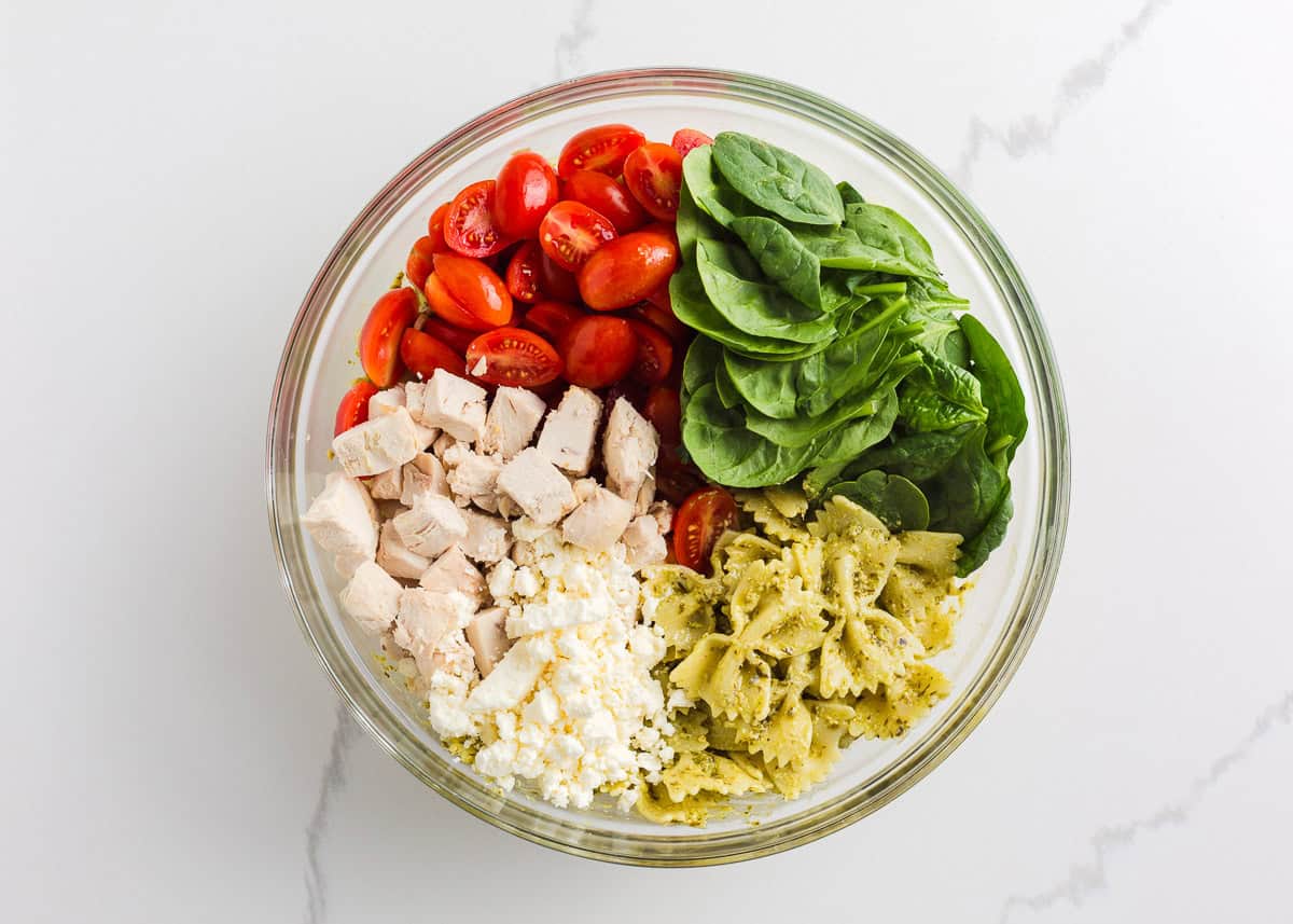 Cooked chicken, halved cherry tomatoes, spinach, feta and pasta tossed with pesto in a bowl.