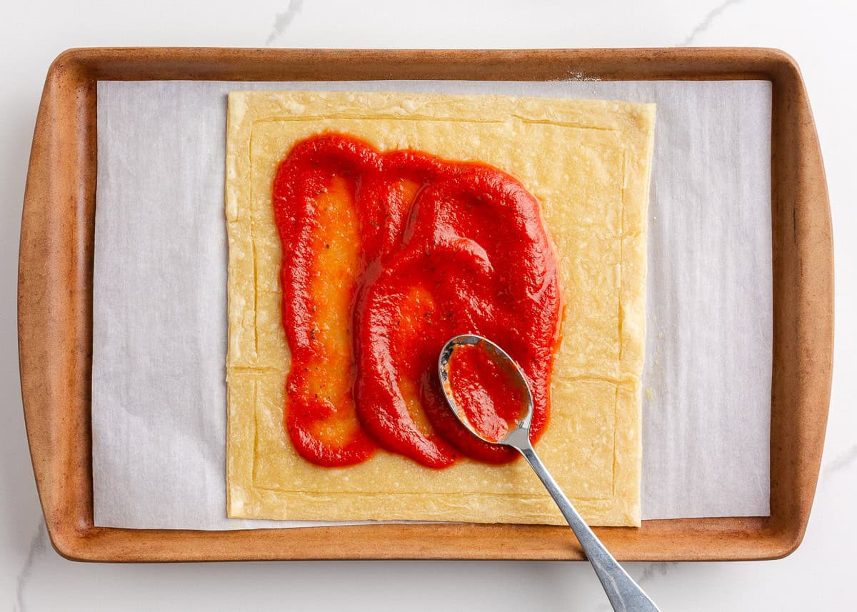 Spreading pizza sauce on puff pastry crust.