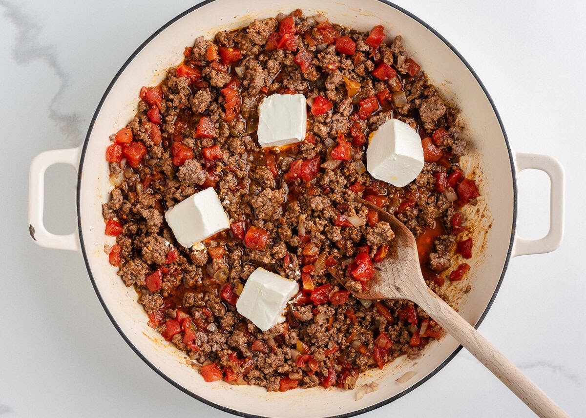 Ground beef in a skillet being mixed with cream cheese, taco seasoning, and diced tomatoes with green chilies.
