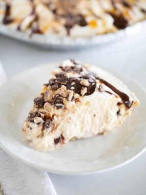 Slice of ice cream pie on a plate with full pie in the background, topped with hot fudge, caramel, and toffee bits.