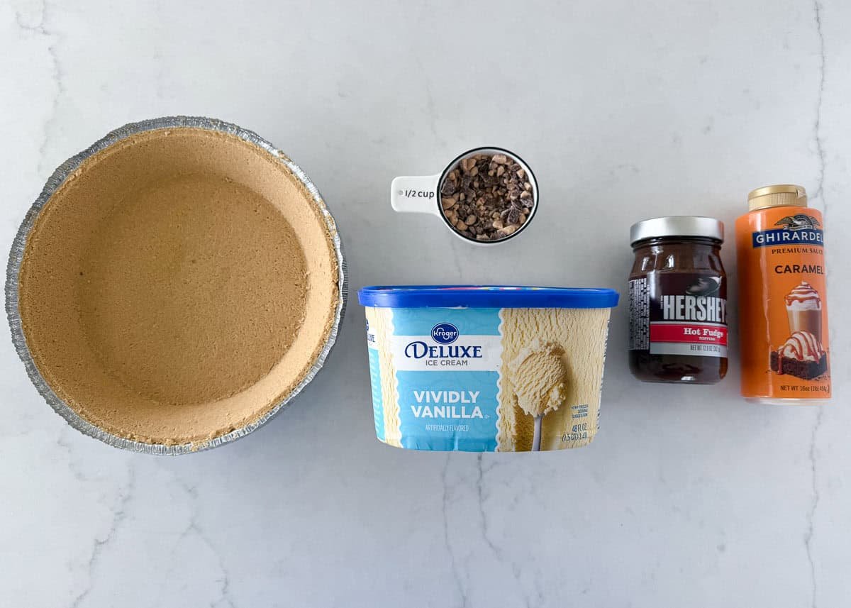 This easy ice cream pie layers vanilla ice cream in a graham cracker crust with hot fudge, caramel, and toffee bits. A no-bake summer dessert favorite with only 5 ingredients! Flat lay of ingredients for ice cream pie including vanilla ice cream, sauces, toffee bits, and graham cracker crust