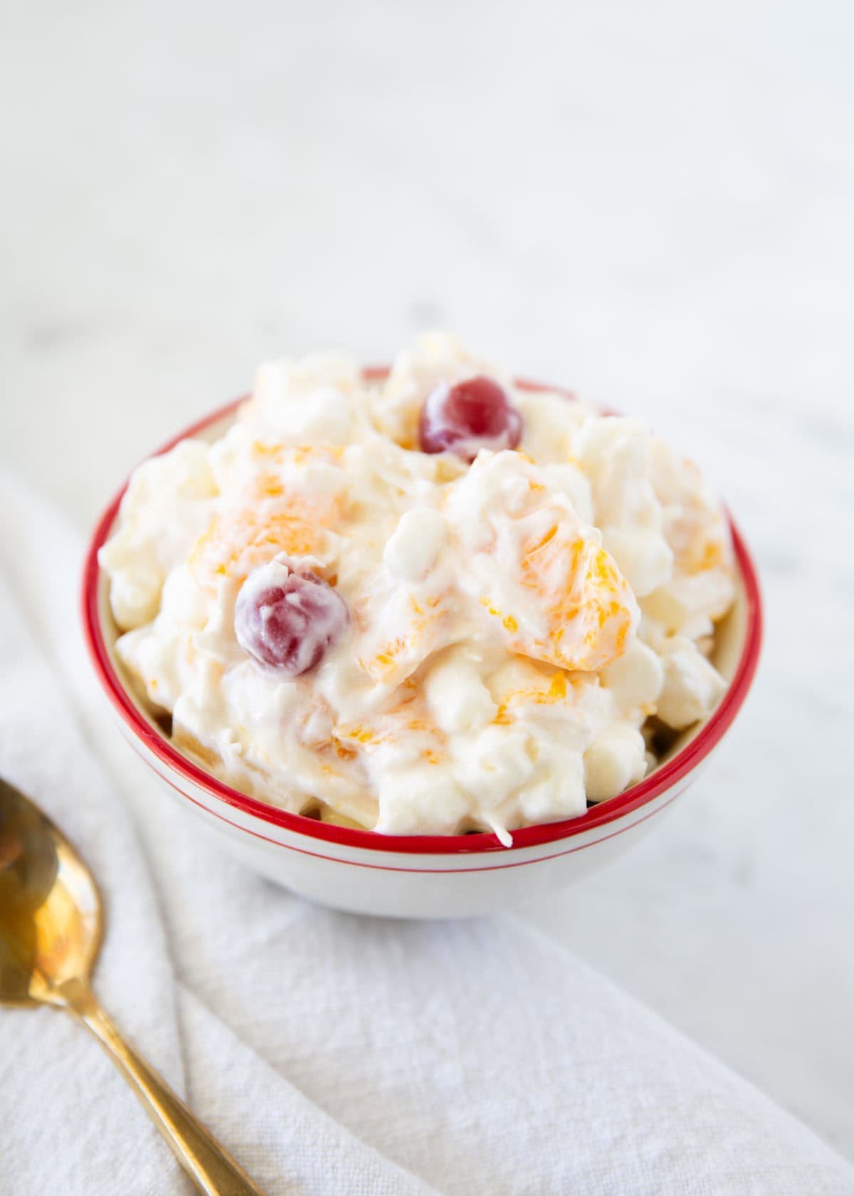 Close-up of 5 cup salad in a white bowl with red trim, showing creamy texture and pieces of fruit and cherries.