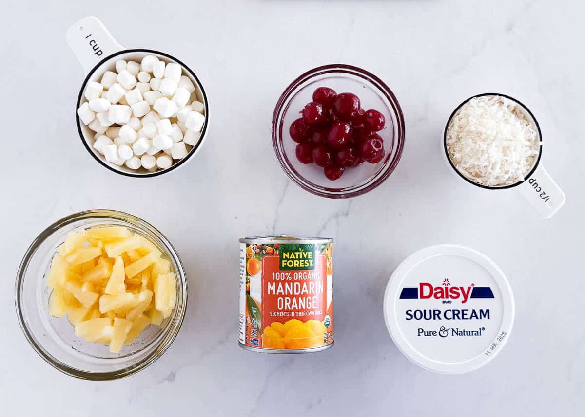 Ingredients for 5 cup salad arranged on a counter, including mini marshmallows, maraschino cherries, shredded coconut, pineapple, canned mandarin oranges, and sour cream.