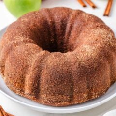 Apple cider donut cake on a white plate on the counter with apples and fresh cinnamon sticks.