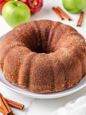 Apple cider donut cake on a white plate on the counter with apples and fresh cinnamon sticks.